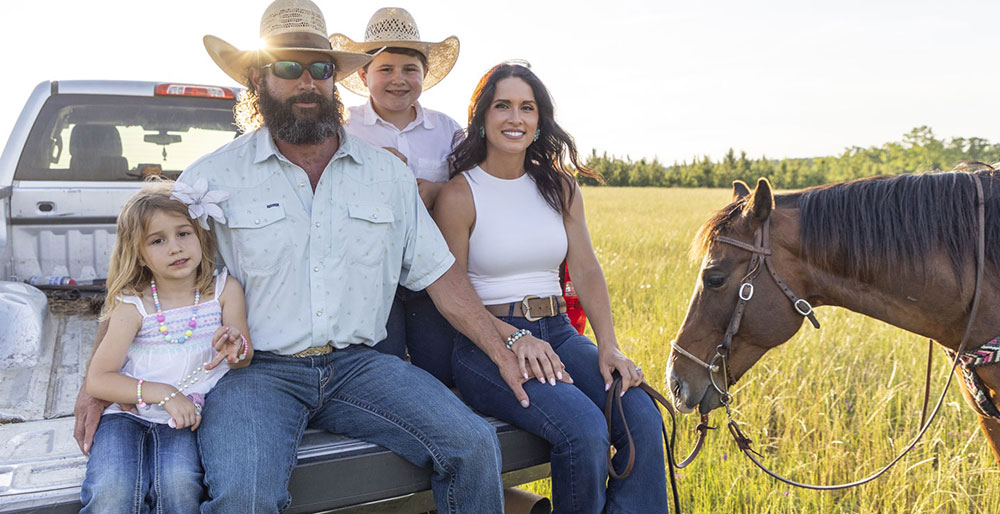 The Smith family sitting in the back of a truck, smiling, while Kip Smith holds the lead rope to a brown horse standing beside the truck. 